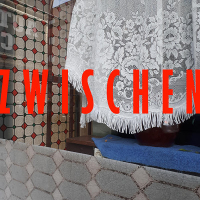 Das Titelbild des Podcasts „Zwischen“ zeigt das Wort in roter Schrift vor einem Foto mit dem Blick in ein Fenster hinein. Im Innenraum sieht man eine Spitzengardine und weiß-rot-grüne Fliesen.