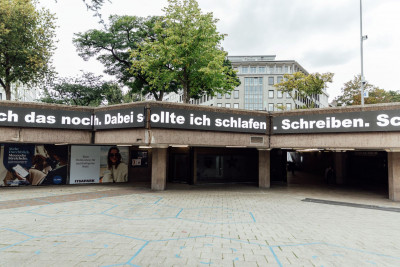 Das Foto zeigt den leeren Ebertplatz in Köln mit einem Betongebäude und einer Überführung. Auf dem Gebäude sind LED Tafeln angebracht, auf denen in weißer Schrift steht: „Dabei sollte ich schlafen. Schreiben. Schreiben.“ Der Bereich darunter ist eine Art überdachter Durchgang mit mehreren Eingängen. Links unten sind Werbeplakate zu sehen. Im Hintergrund sieht man Bäume sowie ein modernes Gebäude mit vielen Fenstern.
